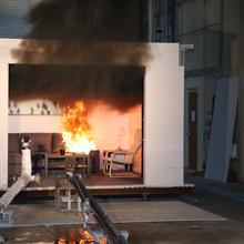 A couch is in flames in a mocked-up room inside a large fire testing facility. 