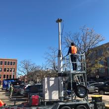 Man in high-vis vest stands on metal platform next to a pole in a parking lot, with a van holding more equipment parked several spots away.