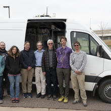Eight adults pose for a photo standing by a white van with the side door open, showing equipment inside. 
