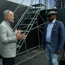 Two men, one wearing virtual reality goggles, standing in a virtual reality testing space