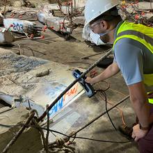 A researcher in safety gear leans over a large piece of concrete and rebar, pointing at it with a scanning device.