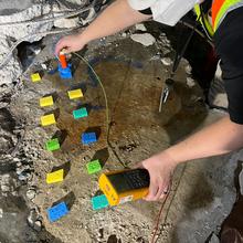 A researcher with a handheld device performs tests with small pieces of colored material lying on a large piece of concrete.