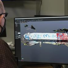 A researcher sits at a computer looking at a model of a broken piece of concrete on the screen. 