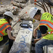 Two people wearing safety gear sit on either side of a concrete column lying on the floor, performing tests on it.