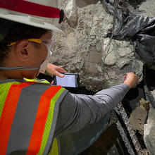 A researcher in safety gear holds a smartphone up to a large piece of concrete and rebar.
