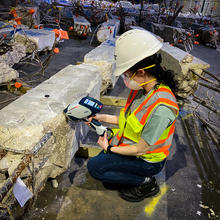 A researcher in safety gear crouches near a large piece of concrete, looking at a handheld device.