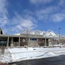 A low office building with a sign saying "NIST" is shown in front of mountains and a blue sky. Snow is on the ground.
