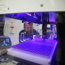 Two scientists watch as a robotic arms moves within a clear plastic box in a laboratory. The scientist on the left isn't there in person, but appears on a computer screen. 