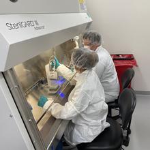 Two researchers in white coveralls and hair coverings use devices to fill vials on a metal counter inside a large transparent hood.