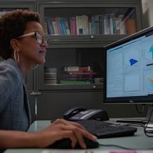 Side view of Danielle Brager at a desk, looking at a monitor, hand on a computer mouse.