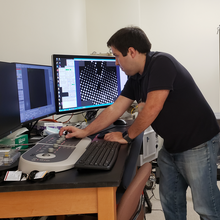 Thomas Cleveland stands at a desk, looking at a computer screen while turning a dial on a control panel. 