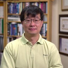 Jun Ye (from the chest up) standing in front of a wall of books and awards. Background is blurred out.