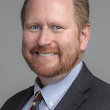 R. Austin Hicklin in a suit and tie. He has red hair, beard, and mustache, blue eye and a smile. 