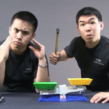 Screenshot from video shows two researchers in NIST T-shirts mugging over a plastic balance. 