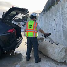 A worker in NIST safety gear stands at the back of an open car trunk, pointing a hand-held device at a concrete column lying broken on the ground near a wall. 