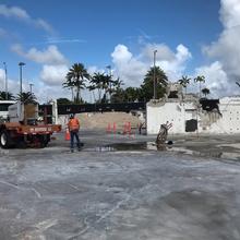 A flatbed truck with equipment on the back sits in a puddled parking lot near the remains of a demolished building foundation, with two figures in safety gear nearby and palm trees in the background. 