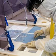 A worker in a white coverall with a yellow apron leans over a counter, using a small instrument on a flat blue material.