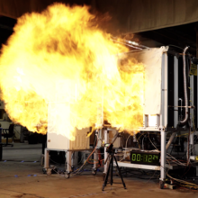 A fireball rushes out of the opening of a small metal compartment in a concrete room. 