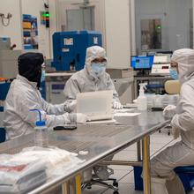 Three people in coveralls sit talking together at a metal table in a high-tech lab.