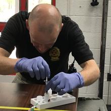 A man in a black shirt labeled "Fire Marshal" is wearing plastic gloves as he bends over a vial in a holder on a table. 