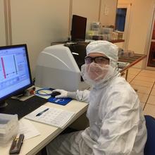 A person in a white coverall and safety glasses poses sitting at a computer in a lab.