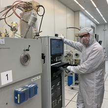 A person in a white coverall and safety glasses poses standing next to a rack of computers and other devices in a lab. 