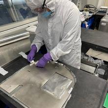 A person in a white coverall and safety glasses bends over a table where she is working on a flat piece of metal. 