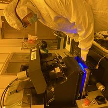 A person in white coveralls and safety glasses looks down at a machine she is using in a lab with orange lighting. 