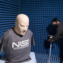 A model head and torso wearing a NIST T-shirt stands on a table in a small room with blue foam on the walls, while a researcher sets up equipment in the background.