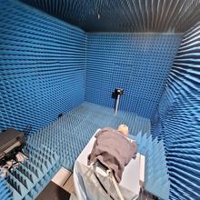 A model head and torso wearing a gray T-shirt lies on a table in a small room with blue foam on the walls. Tubes run from the torso to equipment beneath.