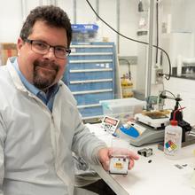 John Kitching poses at a lab table, displaying a chip-scale device in a clear plastic box. 