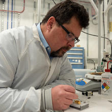 John Kitching leans over a table in the lab, using tweezers to point to part of a chip-scale device in a clear plastic box. 
