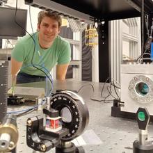 Jacob Davidson poses standing behind a large piece of scientific equipment with wires and lenses.