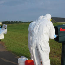 Four figures in coveralls stand outdoors around a mailbox, looking at a clipboard and other items. 