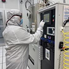 A person in a white coverall and mask stands at a screen on a large piece of equipment in a lab. 