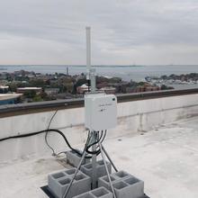 On a rooftop overlooking a waterfront town, an equipment box is attached to a pole anchored with concrete blocks. 