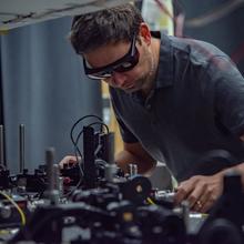 Man wearing safety glasses leans toward a tabletop optical laser setup.