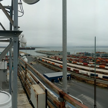 Wide-angle photo shows two people on the left installing equipment on the roof of a building, with a view of a port and rows of shipping containers to the right. 