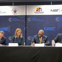 Four officials sit at a table signing documents in front of banners for the NIST National Cybersecurity Center of Excellence.