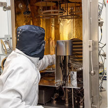 A researcher wearing coveralls reaches into a tall open piece of scientific equipment lined with aluminum foil. 