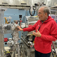 Scott Dewey stands and gestures toward the end of a long metal tube where many connectors and devices are wrapped in aluminum foil. 