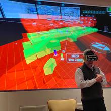 A man wears a virtual reality headset in front of a large projection screen with red and green graphics.