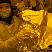 NIST researcher Jessie Zhang, wearing a protective suit, mask, and safety glasses holds up a piece of foil in the lab. 