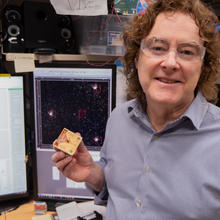NIST's Andrew Wilson holds an ion trap in his lab in Boulder, Colorado. Behind him are screens depicting dots. 