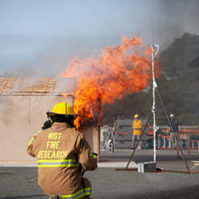 Photo shows a NIST firefighter on standby watching a shed burn next to a target structure representing a wall of a residence during an experiment.