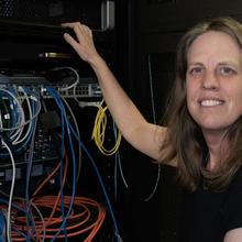 Jeanne Quimby crouches next to a rack of computer equipment with many different colored cord connections. 