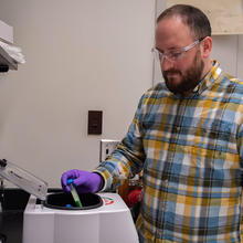Ben Place, wearning safety glasses and gloves, places a vial into a circular opening on a tabletop centrifuge machine. 