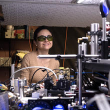 Ana Maria Rey wears dark safety glasses as she stands behind a table covered with optics equipment in the lab. 