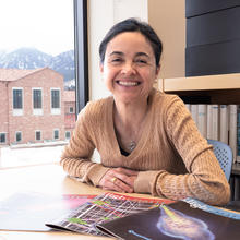 Ana Maria Rey poses at her office desk with physics journals spread out in front of her. Mountains are visible through the window behind her. 