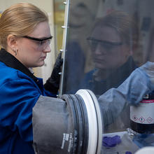 Christine McGinn wears safety glasses as she opens a container inside a clear plastic box in the lab. 